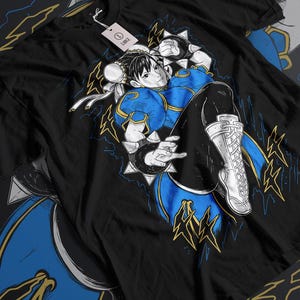 Könnte beinhalten: Ein schwarzes T-Shirt mit einem Grafikdruck einer weiblichen Figur aus einem Videospiel. Die Figur trägt ein blaues und goldenes Outfit und befindet sich in einer Kampfpose. Die Grafik ist von Blitzen und dem Text "Hadoken" umgeben.