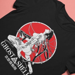Peut inclure: T-shirt noir avec un motif blanc d'une femme avec des fils et un pistolet dans un cercle rouge. Le texte "Ghost in the Shell" et des caractères japonais sont sous le motif.