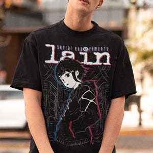 May include: A black t-shirt with a graphic print of an anime character. The graphic is in white, pink, and blue. The text on the shirt reads "serial experiments lain".