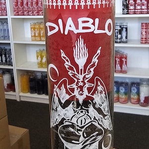 May include: A red glass candle with a black and white image of a winged demon holding a bag of money. The candle is labeled "DIABLO" and "EL PRAS LEVI DYL".