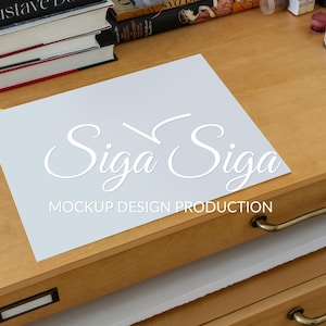 May include: A light blue paper with the text "Siga Siga" in a cursive font and "Mockup Design Production" in a sans-serif font. The paper is on a wooden desk with books and drawers.