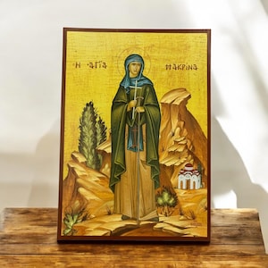 May include: A hand-painted icon of Saint Macrina, a female saint in the Orthodox Christian tradition. The icon depicts her standing in a desert landscape with a cross and staff. The icon is painted on a wooden panel and has a gold background. The text "Η αγια Μακρινα" is written in Greek at the top of the icon.