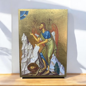 Puede incluir: Icono religioso que representa una figura barbuda con túnicas azules y verdes con alas rojas, sosteniendo un pergamino. El fondo es dorado con texto, y la escena incluye montañas y un pequeño árbol. El icono está sobre una base de madera.