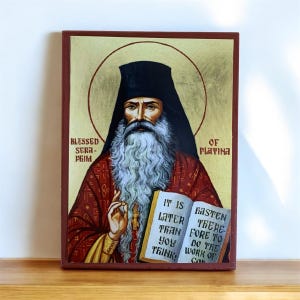 Może przedstawiać: Oprawiona ikona religijna przedstawiająca mężczyznę z długą białą brodą i czarnym nakryciem głowy. Ikona zawiera tekst "Blessed Sera-Phim of Platina" i cytat z książki. Paleta kolorów obejmuje czerwień, złoto i brąz.