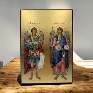 Puede incluir: Un icono religioso rectangular sobre fondo dorado con dos figuras aladas. La figura de la izquierda sostiene una espada y un pergamino, mientras que la de la derecha sostiene un bastón y un orbe. El texto griego dice O ARCHON MIXAHL y O ARCHON GAVRIHL.