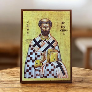 May include: A religious icon featuring a man with a beard, wearing a checkered cloak with black crosses. The icon has gold accents and text in a foreign language. The image is framed in a dark brown wooden border.