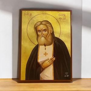 Może przedstawiać: Ikona religijna świętego Serafina z Sarowa, rosyjskiego świętego prawosławnego. Ikona jest namalowana w tradycyjnym stylu ze złotymi akcentami i przedstawia świętego Serafina z długą białą brodą i czarną szatą. Ikona jest oprawiona w ciemną drewnianą ramę.