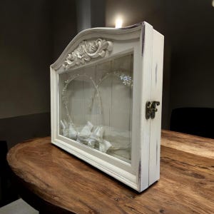 May include: A white, distressed shadow box with a glass front, featuring a heart-shaped wire design with pearls and crystals. The box has a floral top and a metal clasp. Inside are white folded ribbons.