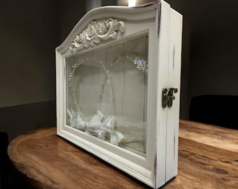 May include: A white, distressed-finish shadow box with a glass front, featuring a heart-shaped wire design adorned with pearls and crystals. The box has a decorative floral top and a metal clasp. Inside are white folded ribbons.