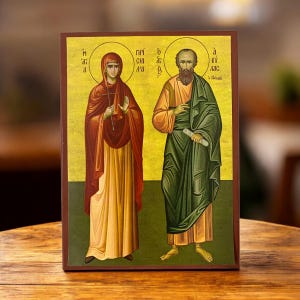Pode incluir: Um ícone religioso com duas figuras com auréolas, vestidas com vestes. A mulher à esquerda usa uma capa vermelha e segura uma cruz. O homem à direita está envolto em verde e segura um pergaminho. O fundo é dourado e a moldura é castanha.