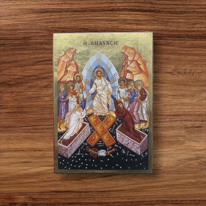May include: A religious icon featuring a central figure in white robes, surrounded by other figures and a golden background. The image includes the Greek text "&Eta; &Alpha;&Nu;&Alpha;&Sigma;&Tau;&Alpha;&Sigma;&Iota;&Sigma;" at the top, with a cross and open tombs in the foreground. The artwork is set against a wooden background.