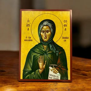Può includere: Un'icona dipinta a mano di Santa Sofia, una donna con una veste verde e un velo blu, che tiene un rotolo. L'icona è su uno sfondo di legno con un'aureola dorata intorno alla testa della santa. Il testo sul rotolo recita "Παράκλησις σας, Ποτί υπέρ ημών".