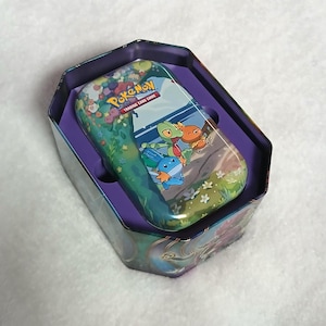 May include: A hexagonal metal Pokémon Trading Card Game collector tin with a purple interior. The lid features a colorful illustration of Treecko, Torchic, and Mudkip in a grassy landscape with flowers, topped with the Pokémon Trading Card Game logo.