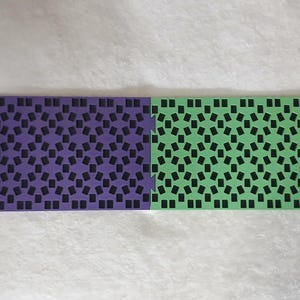 May include: Two interlocking foam floor tiles, one purple and one green, each with a repeating pattern of black rectangles. The tiles are designed to interlock, suggesting a modular flooring system.