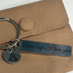 Puede incluir: Un llavero plateado con una etiqueta rectangular grabada con "Drive safe I love you" y una etiqueta circular con la letra "A". El llavero está sobre una funda de fieltro marrón.