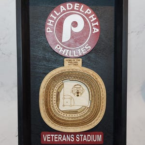 May include: Wooden framed Philadelphia Phillies baseball stadium art. The top features the Phillies logo in red and white. Below is a detailed stadium layout, and a red sign that reads "VETERANS STADIUM".