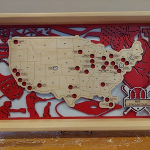 May include: A wooden map of the United States with red baseball stadiums marked on it. The map is framed in a light wood frame. The frame also includes a red baseball glove, a baseball bat, and a baseball with the text "MLB STADIUM TOUR".