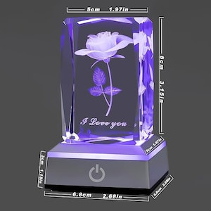 Pode incluir: Um cubo de cristal com uma imagem gravada a laser 3D de uma rosa e as palavras "Eu te amo". O cubo está sobre uma base preta com uma luz roxa e um botão de energia.