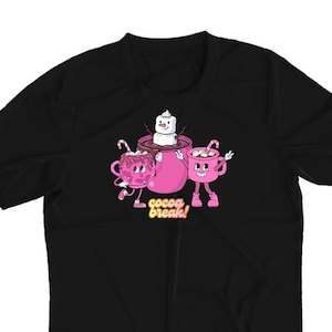May include: Black t-shirt featuring a cartoon graphic of three pink cocoa mugs with faces, marshmallows, and candy canes. The text "cocoa break!" is below the mugs. The shirt is made of a soft material.