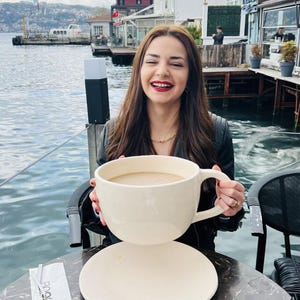 Può includere: Una donna tiene in mano una grande tazza da caffè bianco sporco piena di una bevanda marrone chiaro. La tazza è appoggiata su un piattino abbinato. La scena è all'aperto, con acqua ed edifici sullo sfondo.