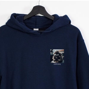 以下が含まれることがあります： 前面に犬の小さな正方形の画像がついたネイビーブルーのフード付きスウェットシャツ。画像の下には「クールストーリー」というテキストがあります。