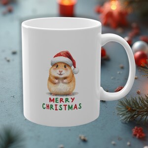 Könnte beinhalten: Ein weißer Keramikbecher mit einer zentralen Illustration eines braunen Hamsters, der eine rot-weiße Weihnachtsmütze trägt. Unter dem Hamster ist der Text Merry Christmas in roten und grünen Buchstaben aufgedruckt. Mit einem seitlichen Henkel.