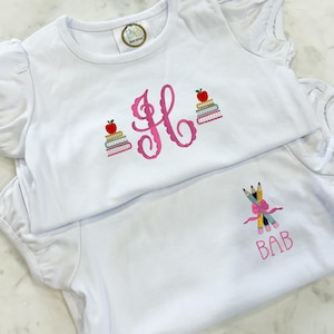 May include: Two white short-sleeved shirts with ruffled sleeves. The top shirt features a large pink embroidered monogram letter H flanked by two stacks of books topped with red apples. The bottom shirt displays three pencils tied with a pink bow and the text BAB.