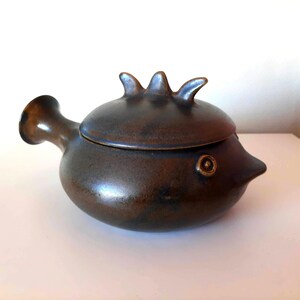 May include: A ceramic, fish-shaped container with a lid. The container is a dark, mottled brown color with a small spout on one side and a decorative fin-like handle on the lid. The eye is a small, circular detail.
