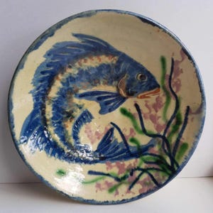 Puede incluir: Plato de cerámica pintado a mano con un diseño de pez azul. El pez está rodeado de plantas acuáticas verdes y rosas sobre un fondo crema. El plato tiene un borde azul y es adecuado para servir alimentos.
