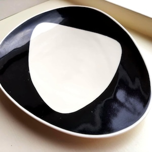 May include: A modern, abstract ceramic dish with a unique shape. The dish features a white interior and a bold, black border that creates a striking contrast. The design is minimalist and stylish, suitable for serving or display.