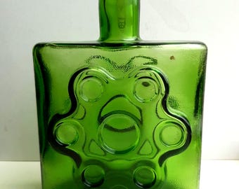 May include: A green glass bottle with a square base and a rounded neck. The bottle features a raised, stylized floral design on the front. The bottle is a vibrant shade of green and appears to be vintage.