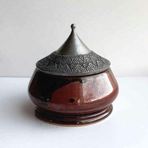 May include: A red and black ceramic jar with a pewter lid. The lid has a pointed top and a decorative design.