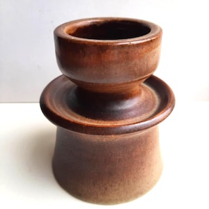 Op de afbeelding: Een handgemaakte, bruine keramische kandelaar met een uniek, gelaagd ontwerp. De kandelaar heeft een brede basis, een centraal platform en een kleine, cirkelvormige bovenkant, ideaal voor een kaars. De kleur is een warm, aards bruin.