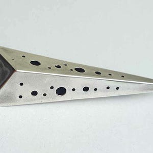 Könnte beinhalten: Ein silberner dreieckiger Anhänger mit einer strukturierten Oberfläche und einem Muster aus kleinen runden Löchern.