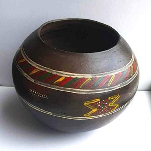 May include: A dark brown clay pot with a wide opening and a rounded bottom. The pot is decorated with white, yellow, red, and green painted designs. The designs include geometric patterns and a stylized symbol.