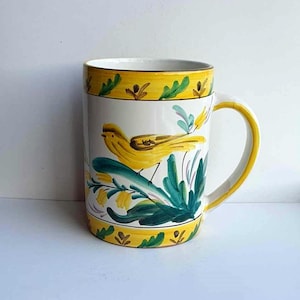 May include: A white ceramic mug with a yellow bird and green foliage painted on it. The mug has a yellow rim and a brown handle.