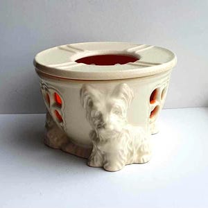 May include: Cream-colored ceramic ashtray with a dog figurine on the front. The ashtray has a round top with a recessed area and a red interior. The sides feature heart-shaped cutouts. A decorative item for the home.