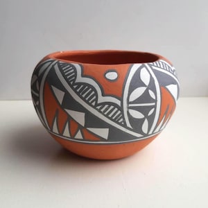 Peut inclure: Un bol rond en céramique peint à la main, avec une base et un intérieur en terre cuite. L'extérieur présente un motif géométrique en gris, blanc et orange. Le bol est probablement utilisé à des fins décoratives ou de service.