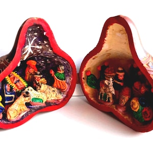 Può includere: Due presepi a forma di zucca. Ogni zucca è scolpita con un interno rosso e una scena della natività dettagliata. L'esterno delle zucche è marrone con disegni dipinti. Le scene della natività includono figure di persone e animali.