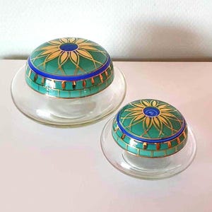 Pode incluir: Dois frascos de vidro com tampa, decorados com desenhos florais verdes e dourados. As tampas têm uma faixa azul e uma borda dourada. Os frascos estão sobre uma superfície branca.