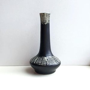 Peut inclure: Un vase en céramique noir avec un col fin et une base bulbeuse. Le vase présente un motif géométrique blanc autour de la base et du bord. Le vase mesure environ 20 cm de haut.