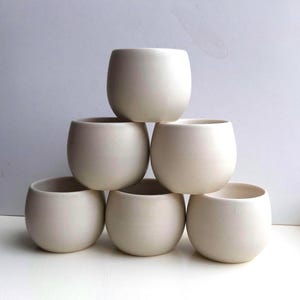 May include: A stack of six small, off-white ceramic cups. The cups have a rounded, egg-like shape with a wide opening. The cups are arranged in a pyramid shape, with one cup on top, two in the middle, and three on the bottom.