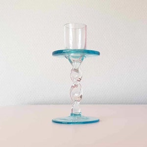May include: A clear glass candlestick with a teal base and top, connected by a twisted stem. The top holds a small glass cup. The candlestick is set against a white background.