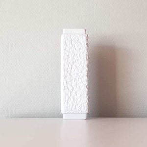 May include: A white ceramic vase with a textured surface. The vase is tall and narrow, with a square base.