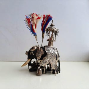 May include: A detailed silver elephant figurine with intricate carvings and a black body. It features a decorative tower on its back, two colorful tassel accents, and a small bell. The elephant has ivory tusks and is a unique decorative item.
