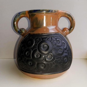May include: A ceramic vase with two handles, featuring a dark gray floral design on the lower half. The vase has a tan body with a blue and green rim. The handles are arched and the vase has a rounded shape.