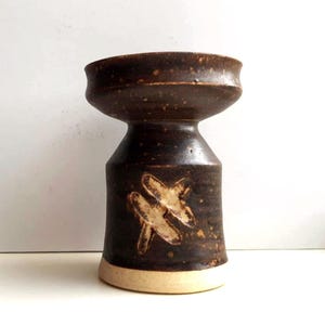 May include: A handmade, dark brown ceramic vase with a unique shape. The vase features a wide, flared top and a cylindrical body with a cream-colored base. Two abstract, cream-colored designs are on the body of the vase.