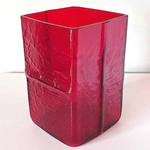 Può includere: Un vaso quadrato in vetro rosso con una superficie testurizzata. Il vaso ha una forma leggermente irregolare e un colore rosso intenso.