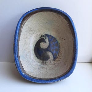 Peut inclure: Bol en céramique ovale avec un bord bleu et un motif central. Le bol présente un intérieur blanc cassé moucheté et un motif en forme d'oiseau bleu foncé et gris. Le bord est bleu vif et le bol a un aspect rustique et fait à la main.