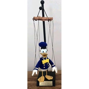May include: A Donald Duck marionette with a blue sailor suit, yellow bow tie, and white gloves. The puppet is suspended by black strings from a wooden control bar. The base is black with gold lettering.
